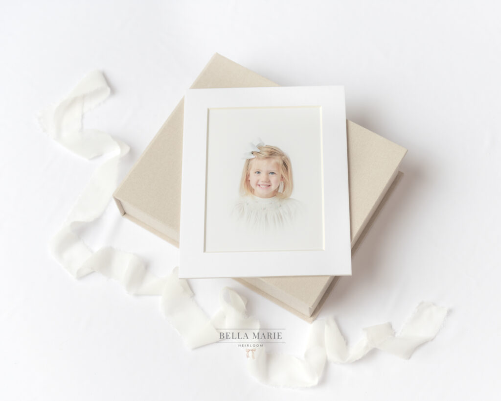 Flat lay of linen box with matted white portrait of a blind child in a white heirloom dress.