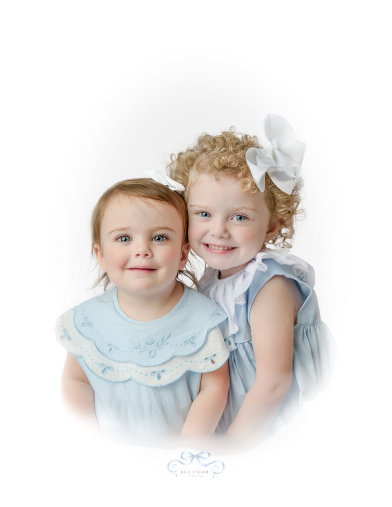 Two little blue eyed girls, one age 4 and one 12 month baby sitting close together, wearing blue heirloom dresses  and smiling for heirloom portrait.   Background is white.