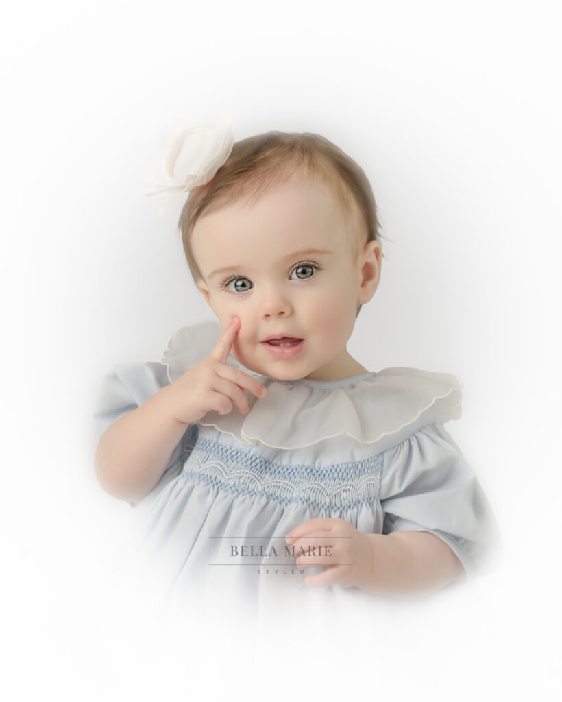 baby girl in a vintage blue dress with large ruffle white collar, blue eyes and brown hair, with finger on her cheek.  White bow in hair and white vignette background.