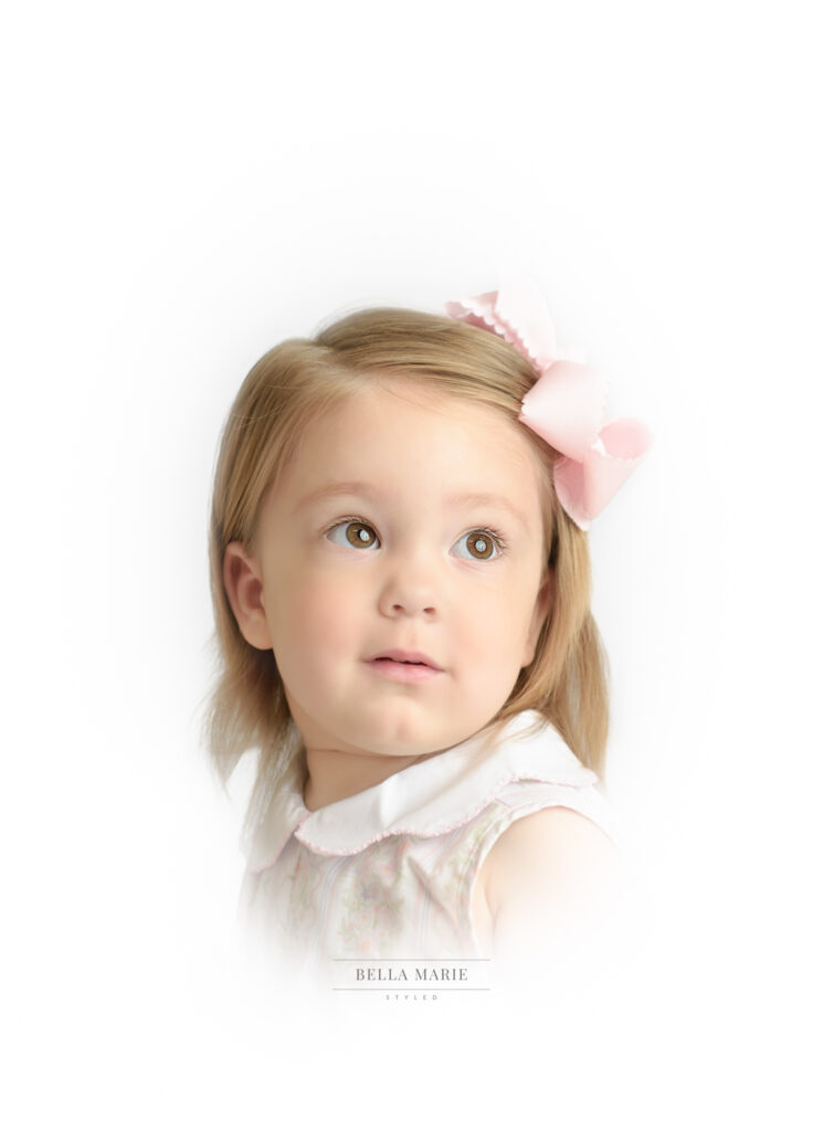 Little girl with sandy blonde hair and pink bow looking up for her portrait.   Her eyes are brown and she is wearing a white Peter Pan Collar shirt.