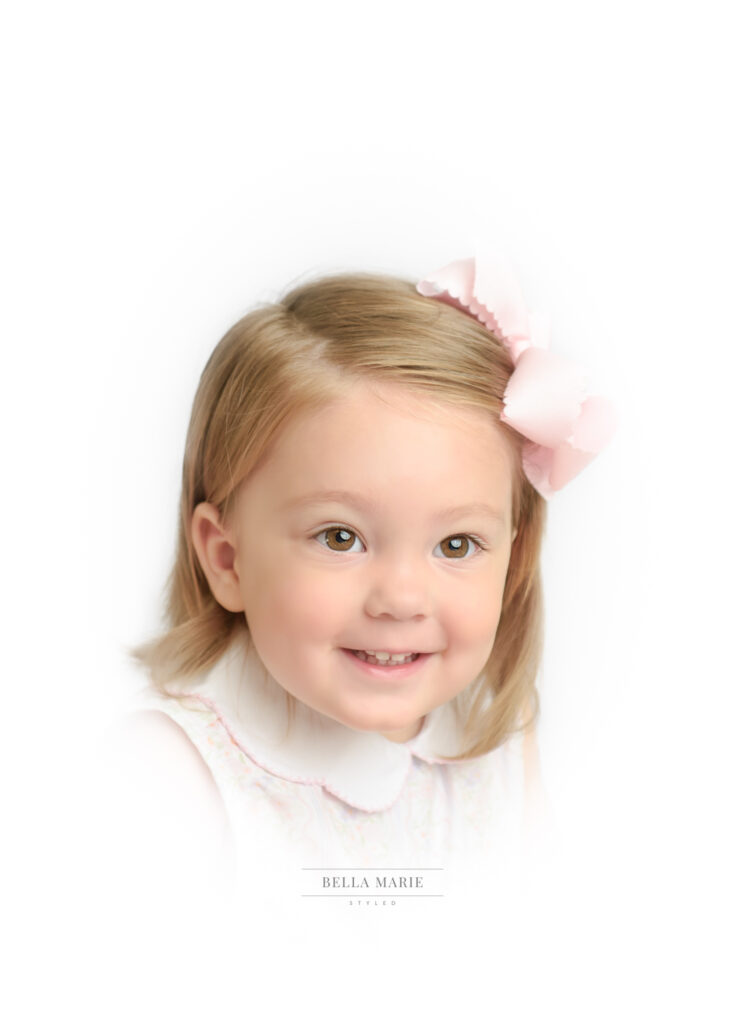 Little girl age 2 with blonde hair and brown eyes smiling with a pink bow in her hair.  Headshot from shoulders up and a white vignette.