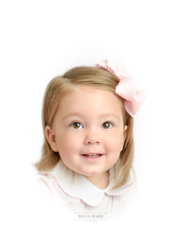 Toddler girl with pink bow in her blonde hair, smiling for camera, headshot with white background.